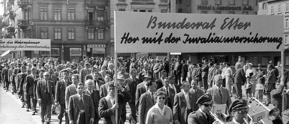 1. Mai-Umzug: Marsch mit Musizierenden und Banner "Bundesrat Etter, her mit der Invalidenversicherung", 1957, Quellennachweis: Schweizerisches Sozialarchiv, Zürich.